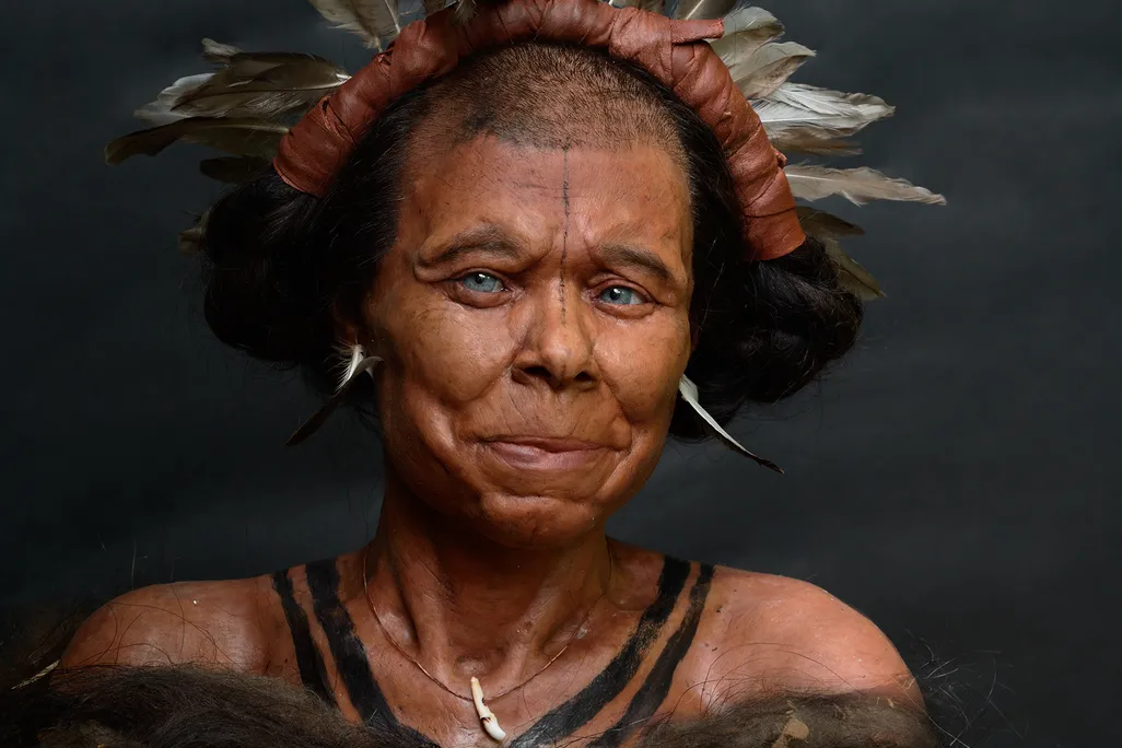 a woman with very short hair, a feather headdress, blue eyes and feather earrings smiles slightly. The image is from her shoulders up