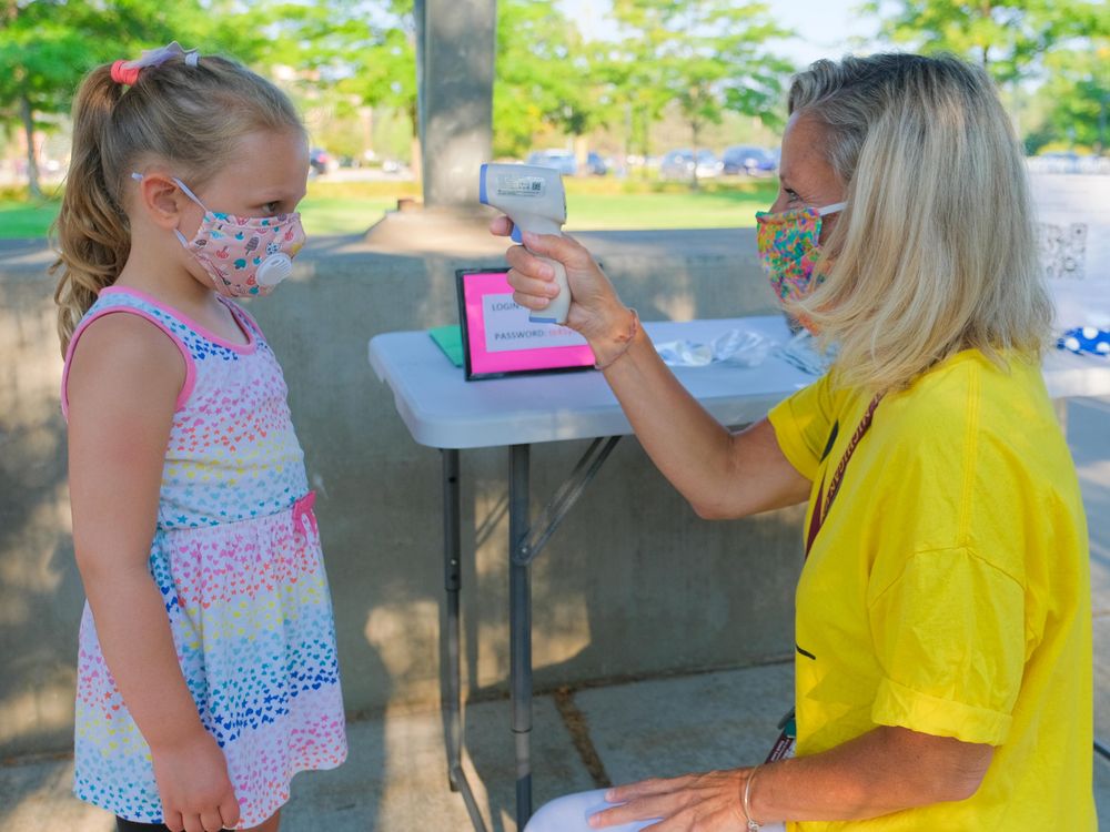 A young girl wearing a colorful cloth mask (left) gets her temperature read by a woman at the Childhood Development and Learning Lab at Central Michigan University