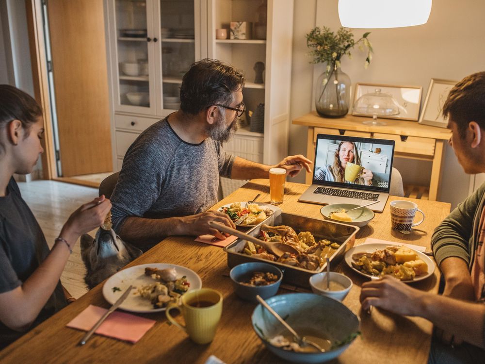 Three people sit around a table for Thanksgiving dinner while having a video call with a woman holding a mug. 