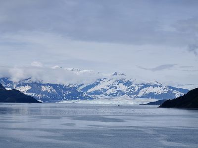 A landscape shot featuring a cloudy gray sky, snow-covered mountains in the distance above a turquoise expanse of glacial ice on the horizon and a dark blue expanse of water in the foreground.