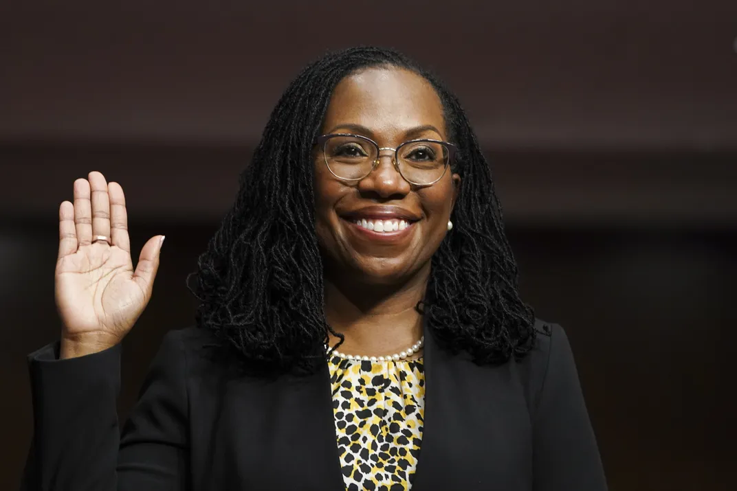 A Black woman with shoulder length braids raises her right hand and smiles at a swearing-in ceremony. She wears a black blazer, a yellow top with black flowers and glasses.