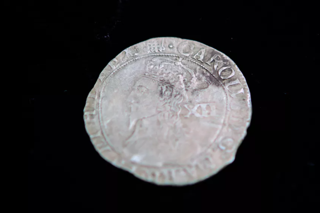 A silver coin with the face of a King and large capital letters going around in a circle; uneven edges