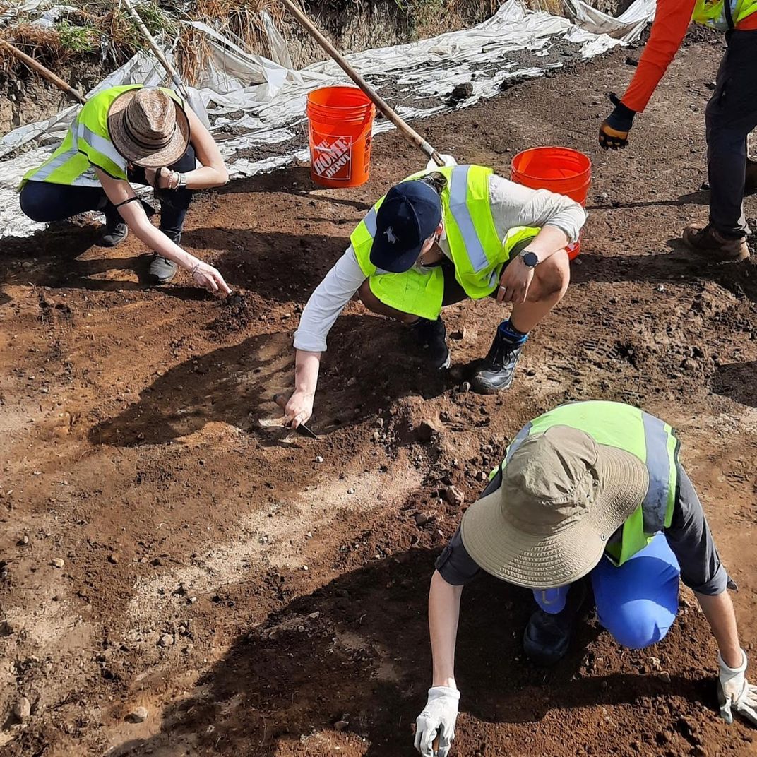 Three people with pale skin, clad in reflective vests, kneel in the brown dirt, scraping carefully at the surface 
