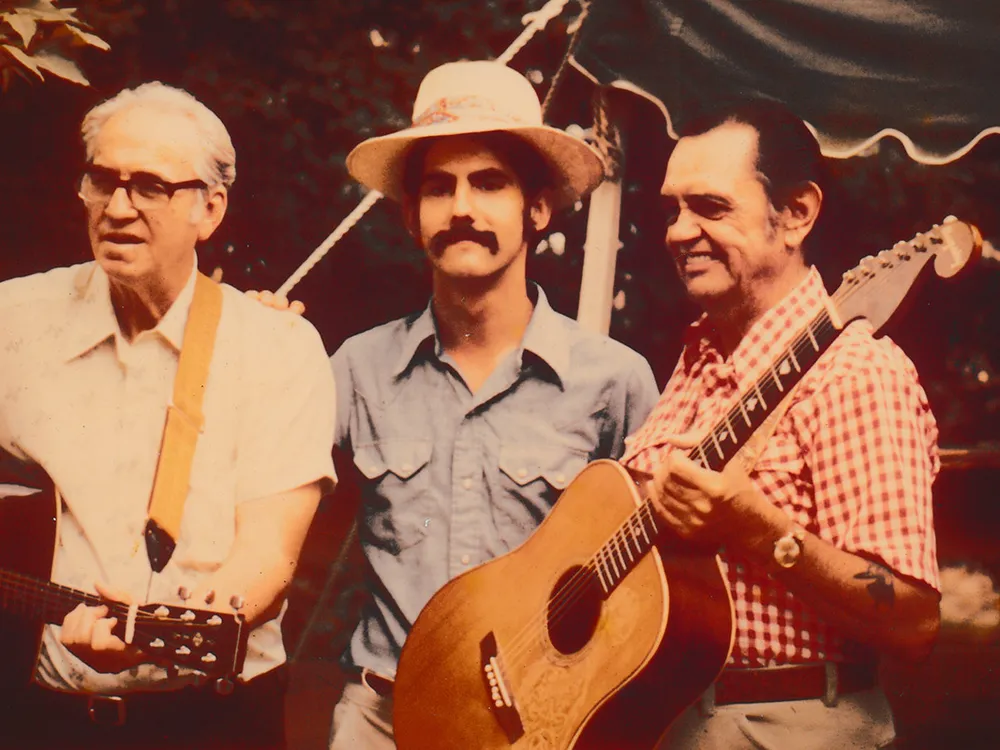 Three men, two holding acoustic guitars, pose in front of a festival tent.