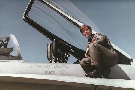 A young woman wearing a U.S. Navy olive-green flightsuit and black combat boots perches next to the cockpit of her F/A-18C miltary jet. "Lt Sharon Cummins" is stenciled onto the side of the airplane.