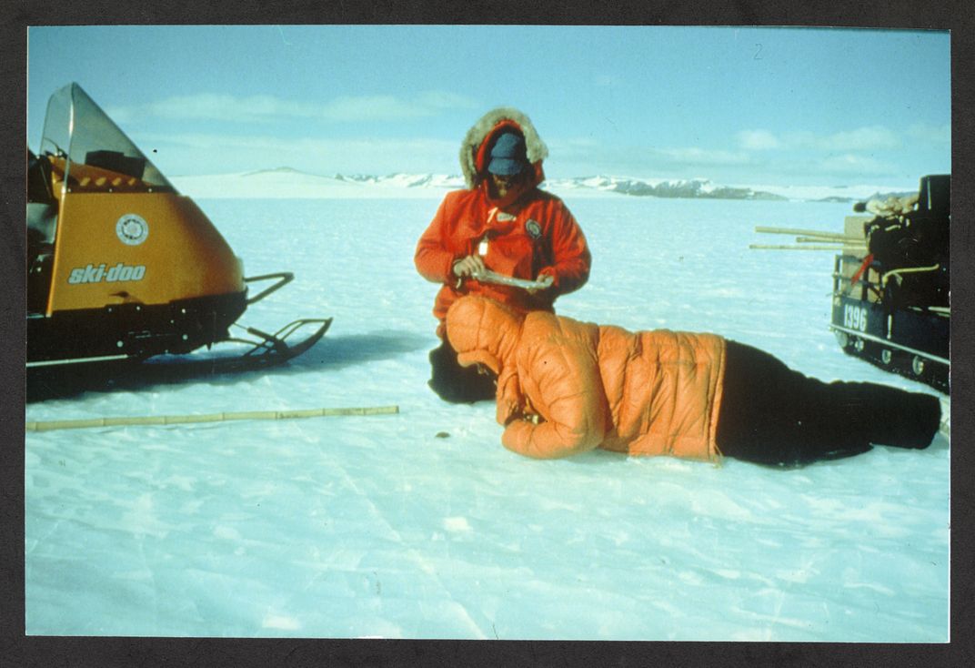 Two figures on glacial ice wearing snow gear. One laying down looking at something on the ground, the other kneeling and writing.