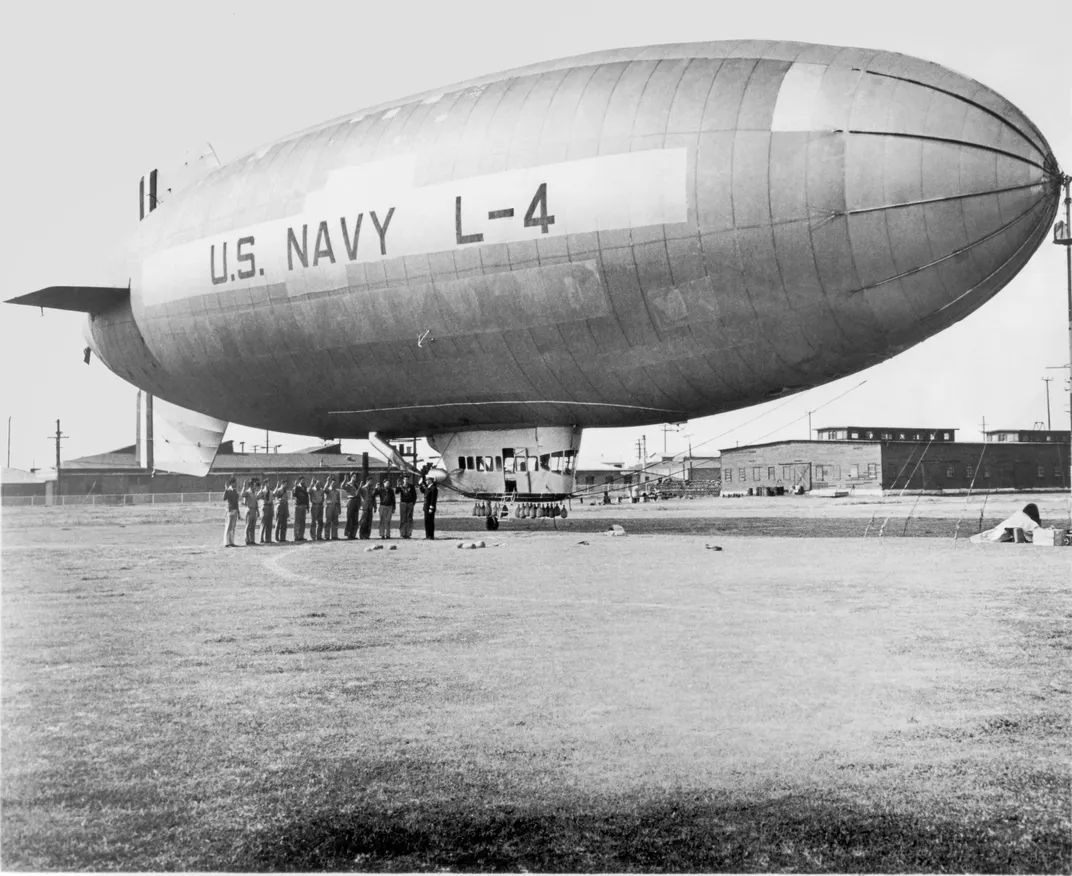 Black and white photo of a blimp floating in the air with the words U.S. Navy L-4 and a group of people standing on the ground in front of it