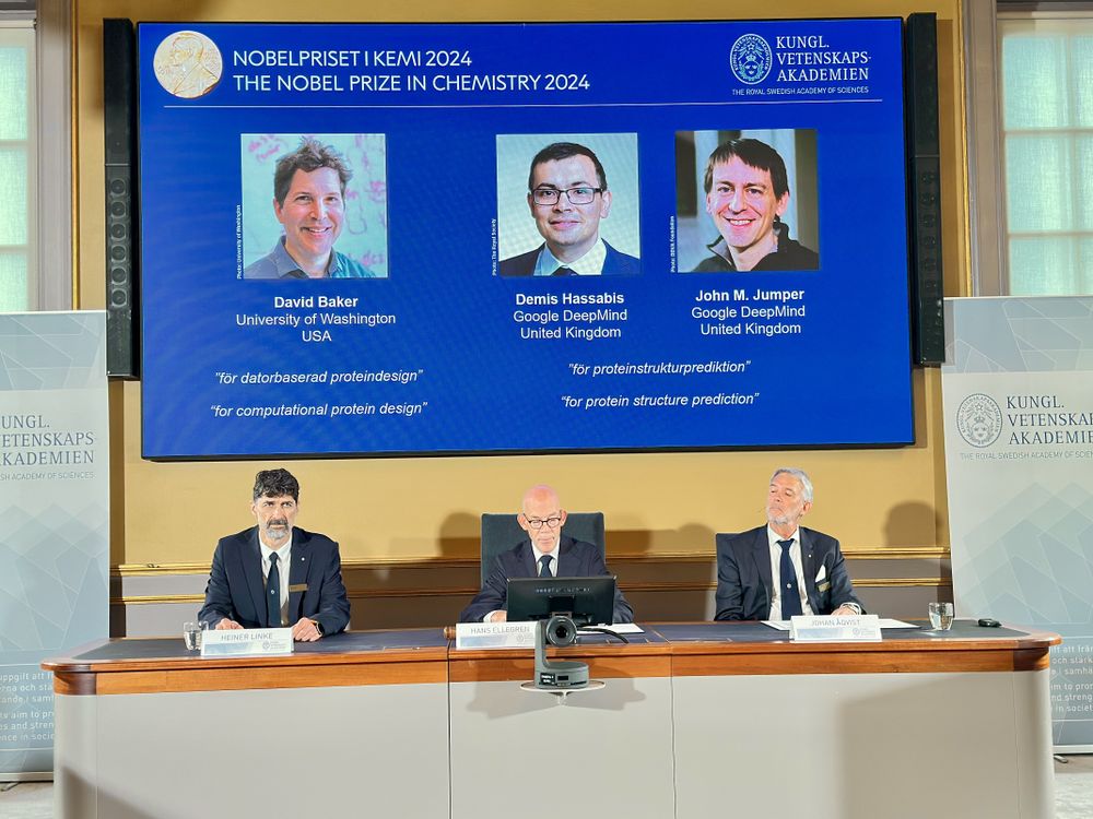 a slide showing the three Nobel winners is projected above three men sitting at a table