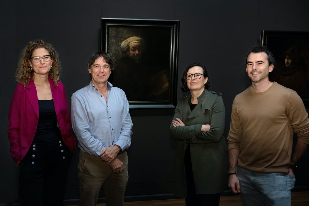 Four people stand next to a portrait of an elderly man in a dark gallery
