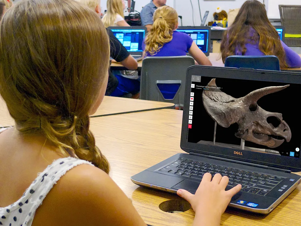 Girl in a classroom using a laptop computer that shows a dinosaur skull on its screen. Several other students work at desks with laptops in the background.