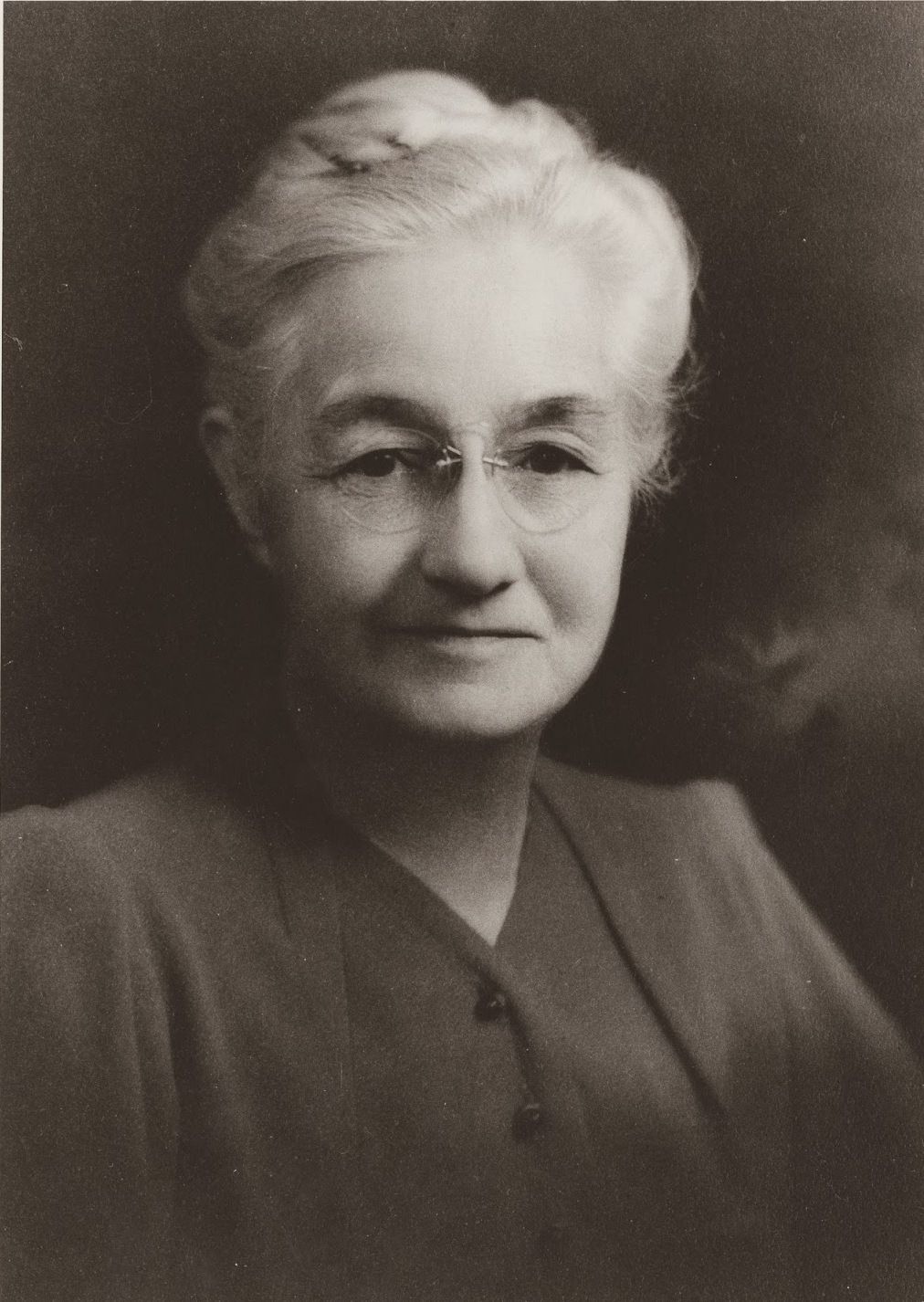 An older women is shown in a traditional portrait style. Her hair is white and up in a bun. She is also wearing glasses.