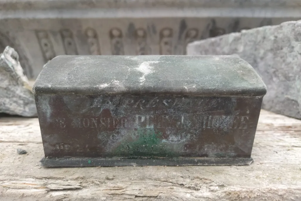 A square metal casket that is rusting and has gone green in some places, close-up, with some inscriptions that read "The heart of Pierre David was solemnly placed in the monument on 25 June 1883"