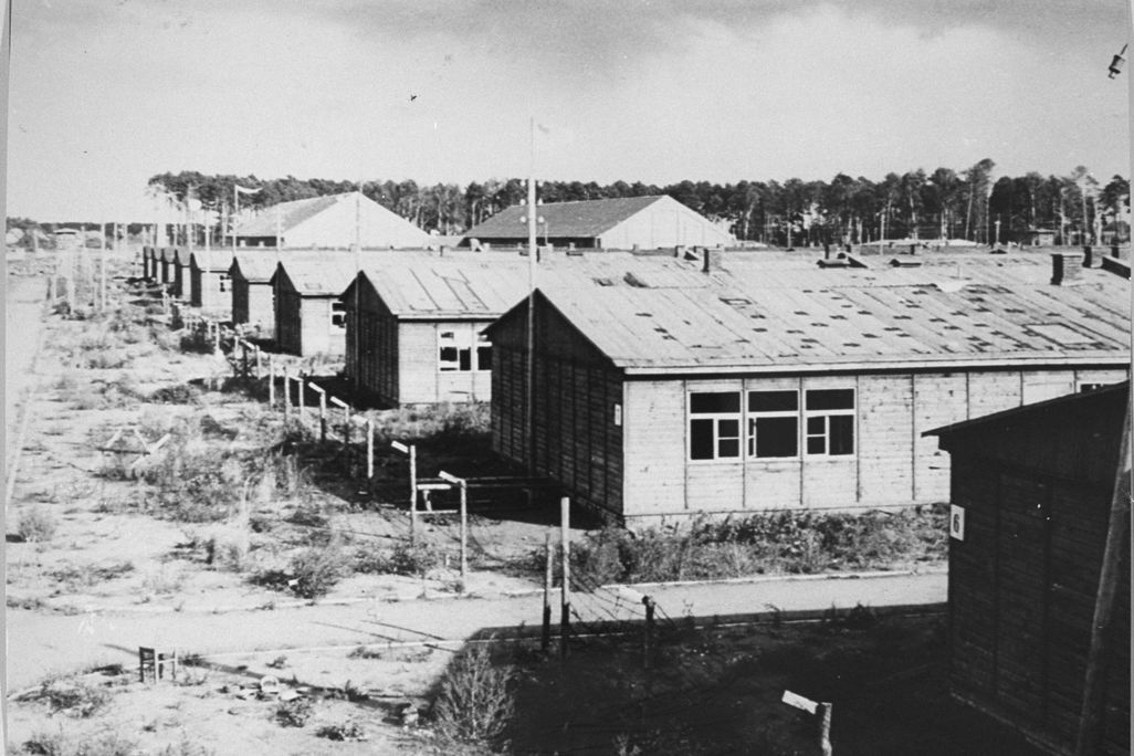 A black and white image of low rectangular buildings with peaked roofs, grassy dirt ground, black windows and shadows