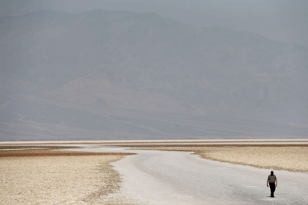 A large expanse of flat, sandy ground, with the faint outline of a hill rising in the distance; one lone traveler is walking in the lower righthand corner of the frame, looking very small compared to the landscape