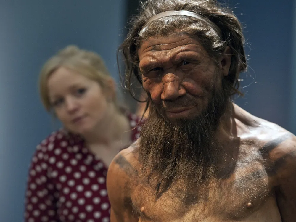 a woman stands behind a model of a neanderthal man, looking over his shoulder