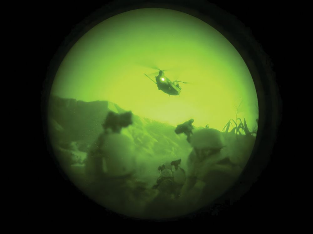 A grainy, green-tinted, circular view through a night vision scope shows a helicopter flying over a dark landscape at night.