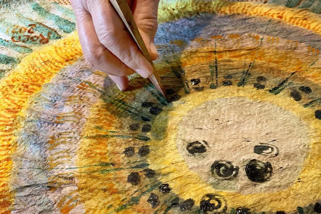 Close-up on a hand holding a wooden painting utensil, sharpened to a point, painting black dots on a bark cloth artwork in yellow, purple, orange, green, and black.