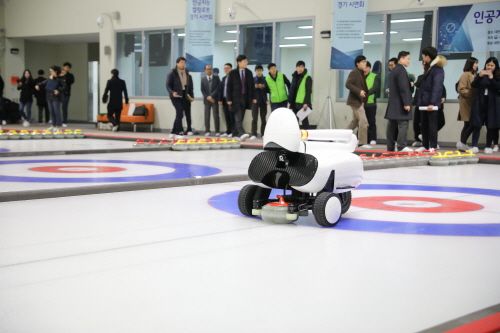 A white robot with a teardrop-shaped "head" appears to crouch down, rolling on wheels, as it pushes a round curling stone forward over ice; many onlookers watching