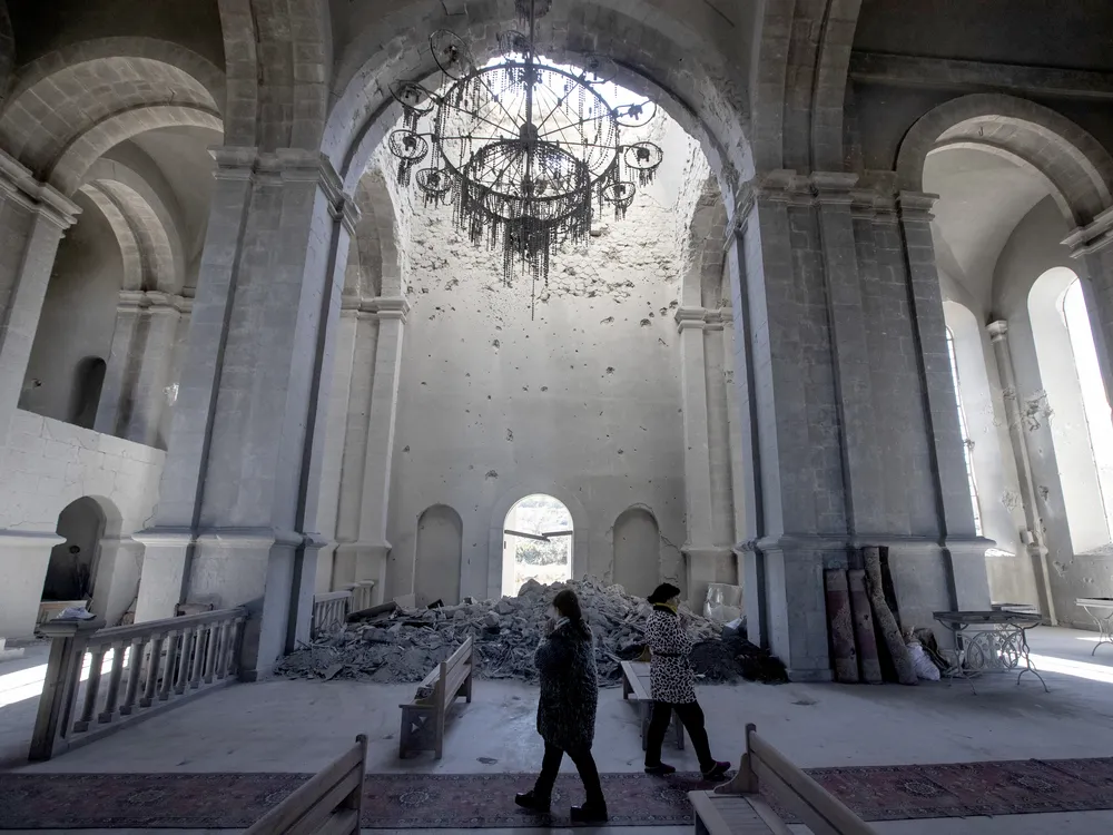 Two people walk, cast in shadows, underneath the tall white arches of a cathedral; behind them, a pile of rubble blocks a doorway and a broken chandelier hangs from the ceiling
