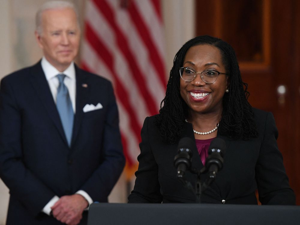 Judge Jackson stands at a podium and smiles, wearing glasses, a string of pearls, a purple top and a black blazer. In the background, President Biden smiles and stands in front of an American flag