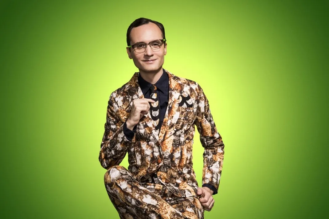 Native filmmaker Joey Clift against a green background sitting with one leg crossed at the ankle over the opposite knee, he wears a suit decorated with faces of cats
