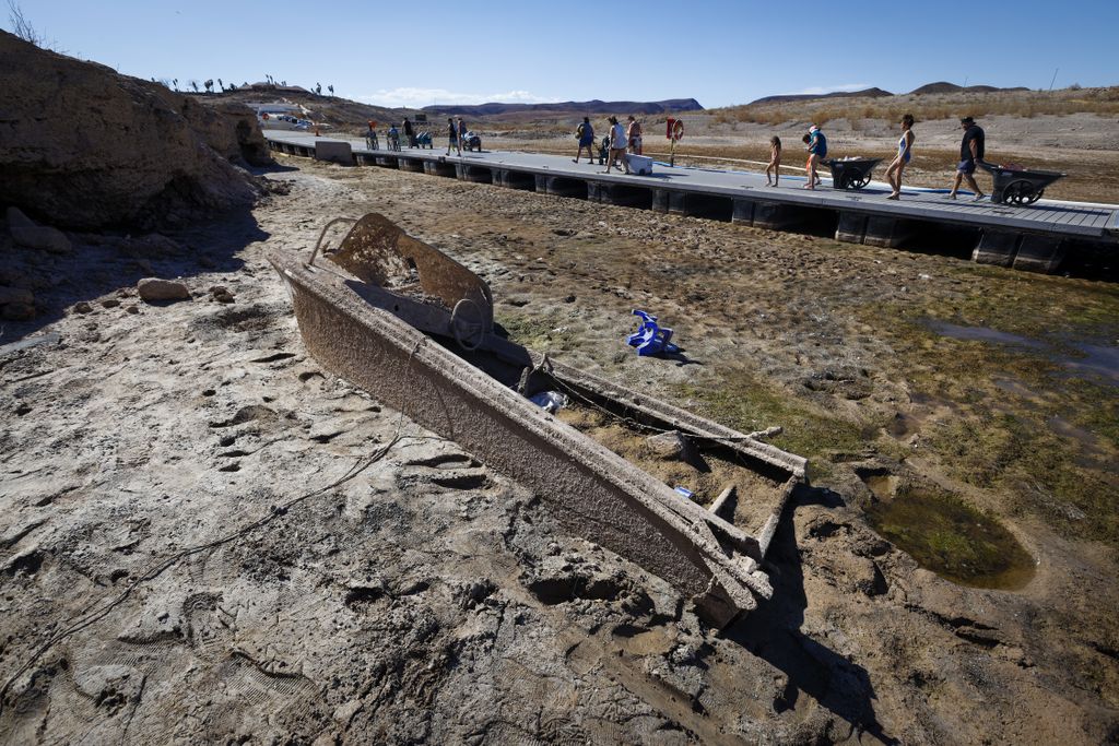 People walk on a makeshift boardwalk across dry land behind a disintegrating, muddy boat