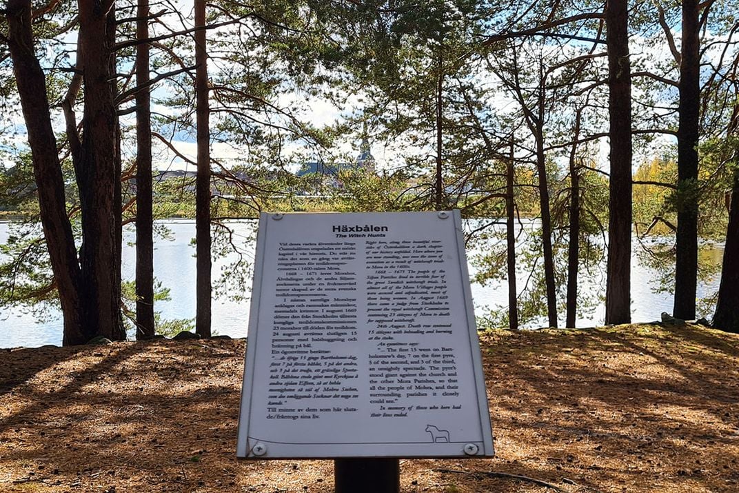 A commemorative sign, written in Swedish, at a seemingly idyllic scene of trees along a river's edge.