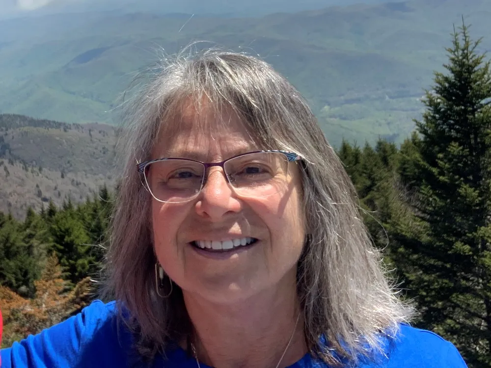 Head and shoulders photo of Denise Breitburg, a woman with glasses and shoulder-length gray hair, with forested mountains in the background.