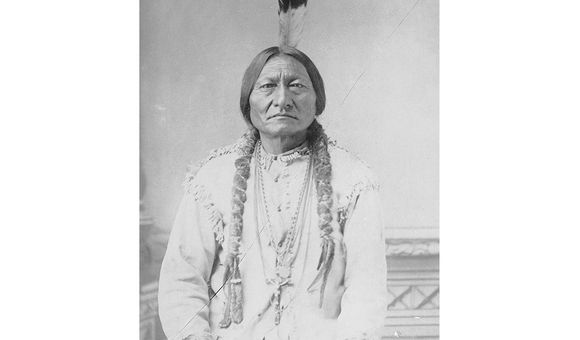 Historic black and white photograph of Sitting Bull sitting down with a feather in his hair and holding a pipe.