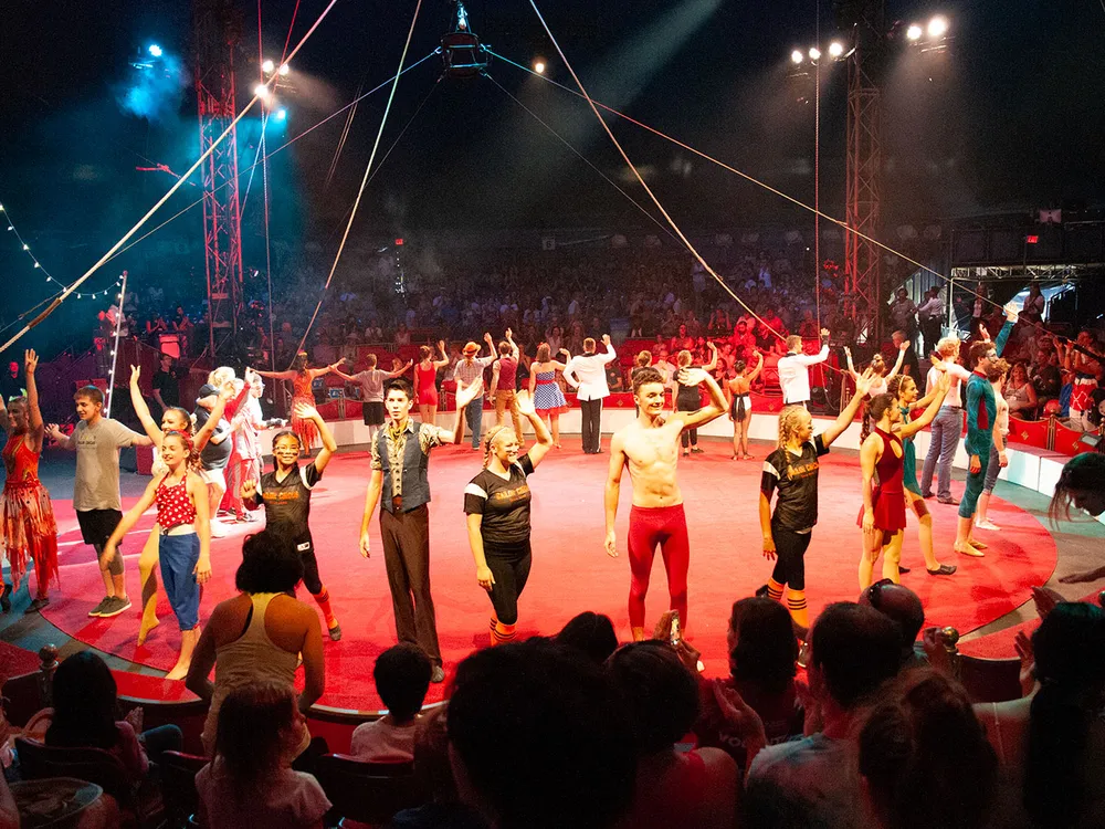 Performers wave, forming a ring around a red, circular stage, with a packed audience and rigging hanging above.