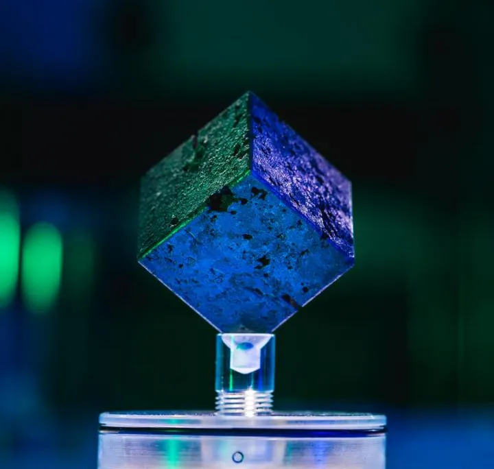 A blue and green image of a small metal cube resting on a display, with colored lights in the background
