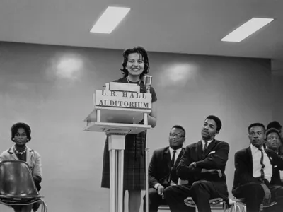 A black-and-white image of Diane Nash at the podium with men and a woman sitting on either side of her in chairs. Nash stands in the center of the image behind the podium labeled [L.R. HALL/ AUDITORIUM] speaking into the microphone.