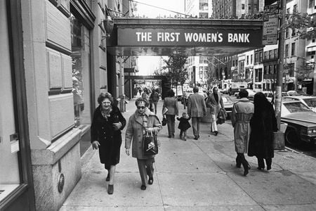Candid black and white photo of people walking down the street. Two women face the camera in the foreground under an awning with the words “The First Women’s Bank” on it.