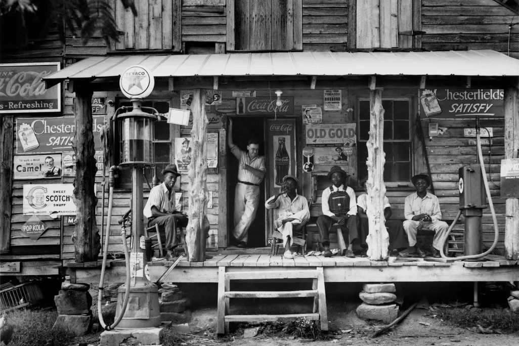 A group of people sit on the porch of a general store, surrounded by signs for products like Coca Cola and Chesterfield's cigarretes; most look at the camera
