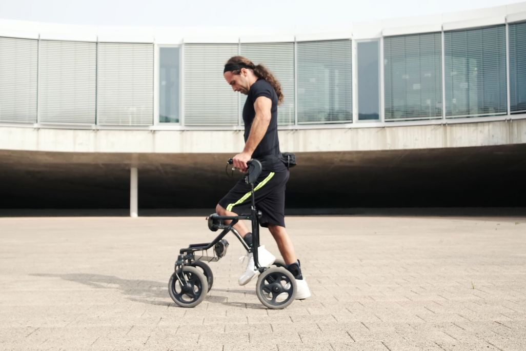 A man with long hair walks with the help of a walker on a concrete surface