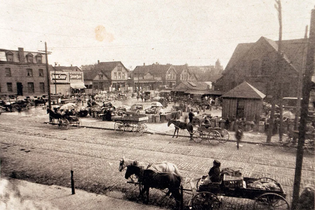 Historic black and white photograph of a busy market square with numerous horse-drawn carriages and pedestrians, featuring cobblestone streets and vintage architecture.