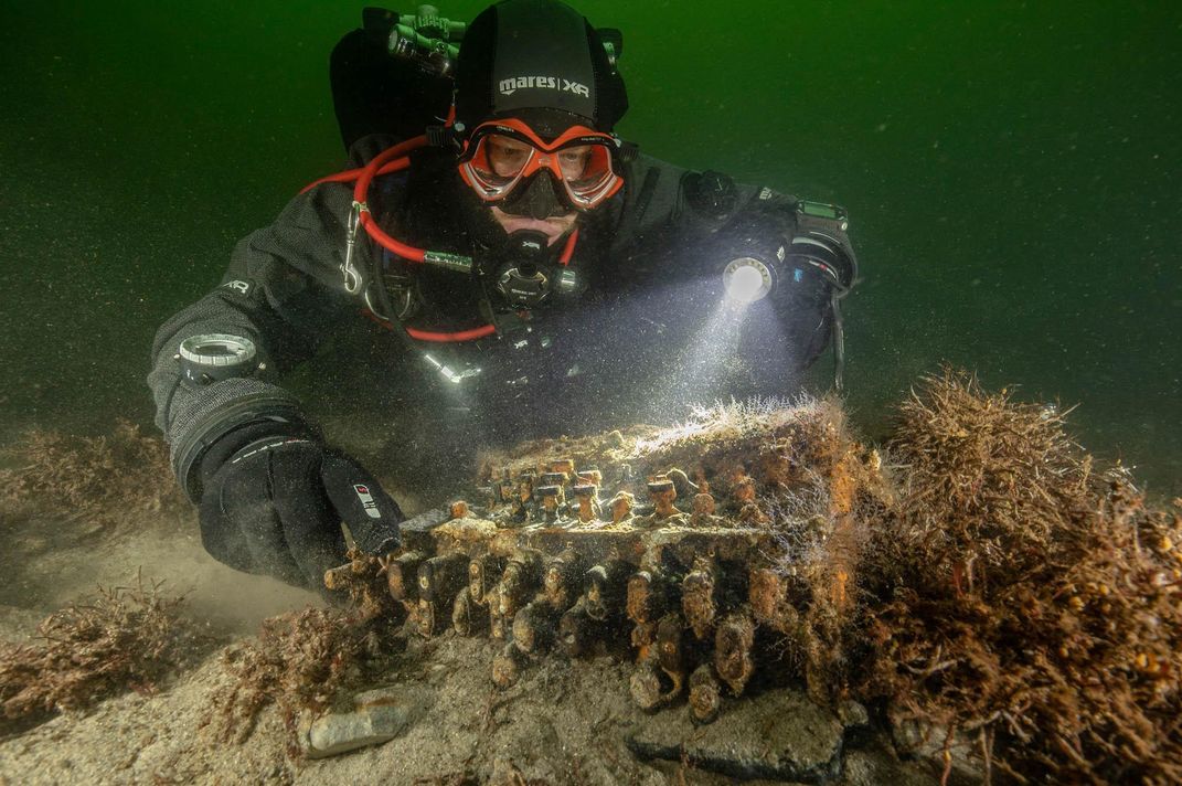 A person wearing scuba equipment and red goggles floats in murky green water and shines a flashlight above an item on the ocean floor, overgrown with algae but resembling a typewriter