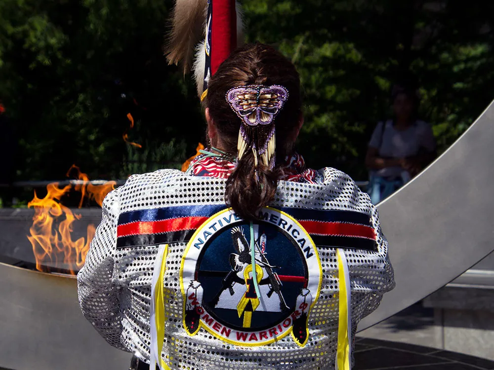 From behind, a woman holds up a flag in front of a memorial monument with a central flame. Her brown hair is braided with a purple butterfly barrette, and she wears white regalia with silver sequins and ribbons across the shoulders.