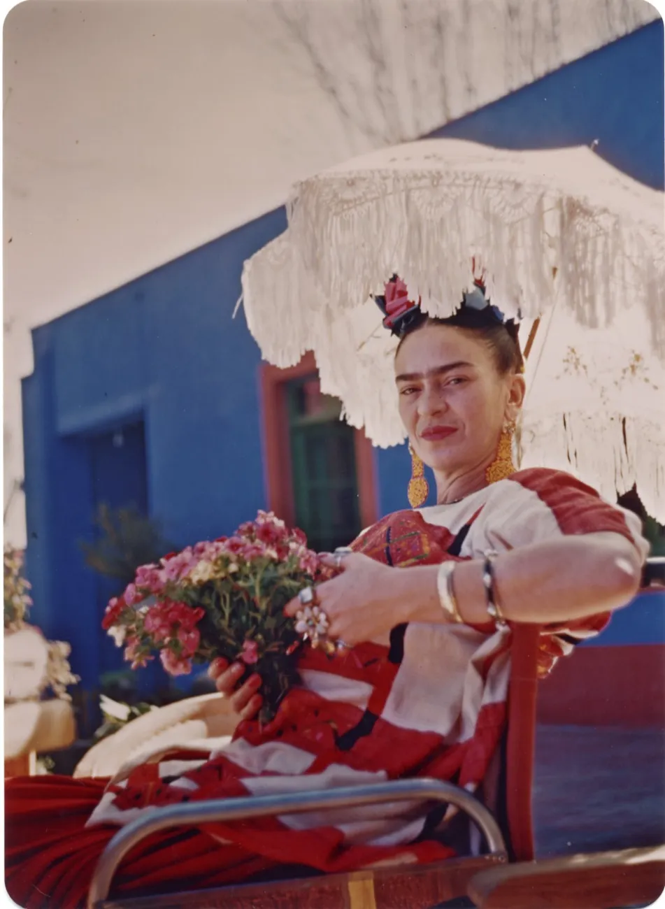 Kahlo holds a bouquet of flowers, wearing a flower crown and red-and-white dress, sitting outside underneath a white parasol in front of her bright blue house