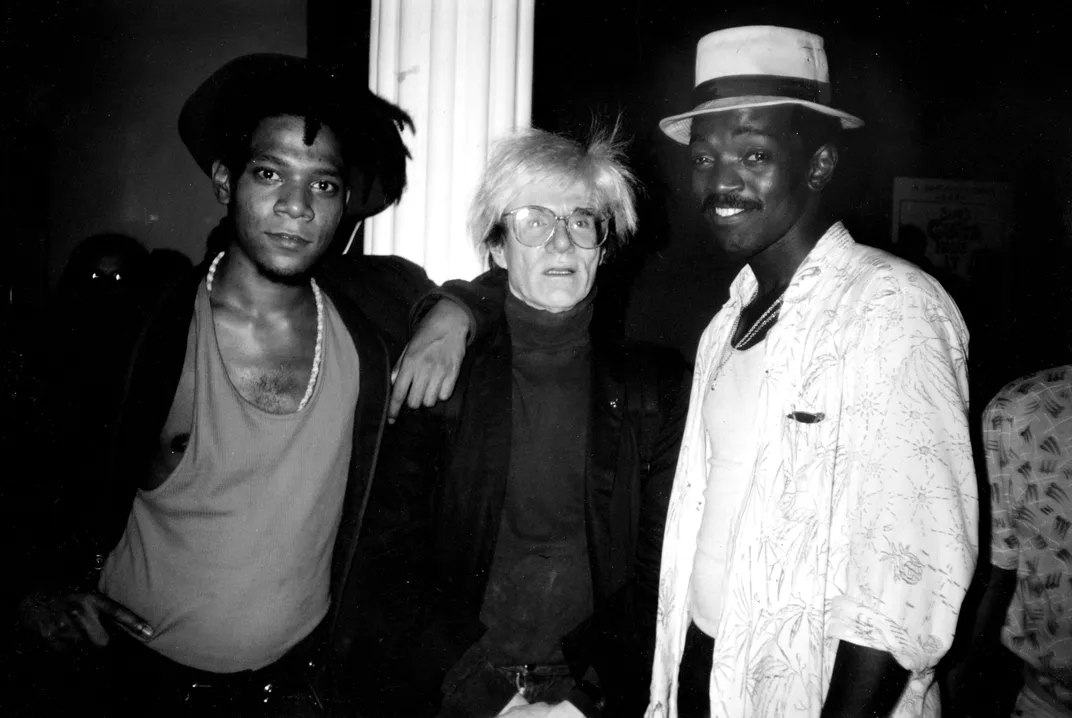 A black and white image of three men, left to right: a young Black man in a deep V-neck, a white man with glasses in a turtleneck and suit, and a Black man in a white shirt and fedora