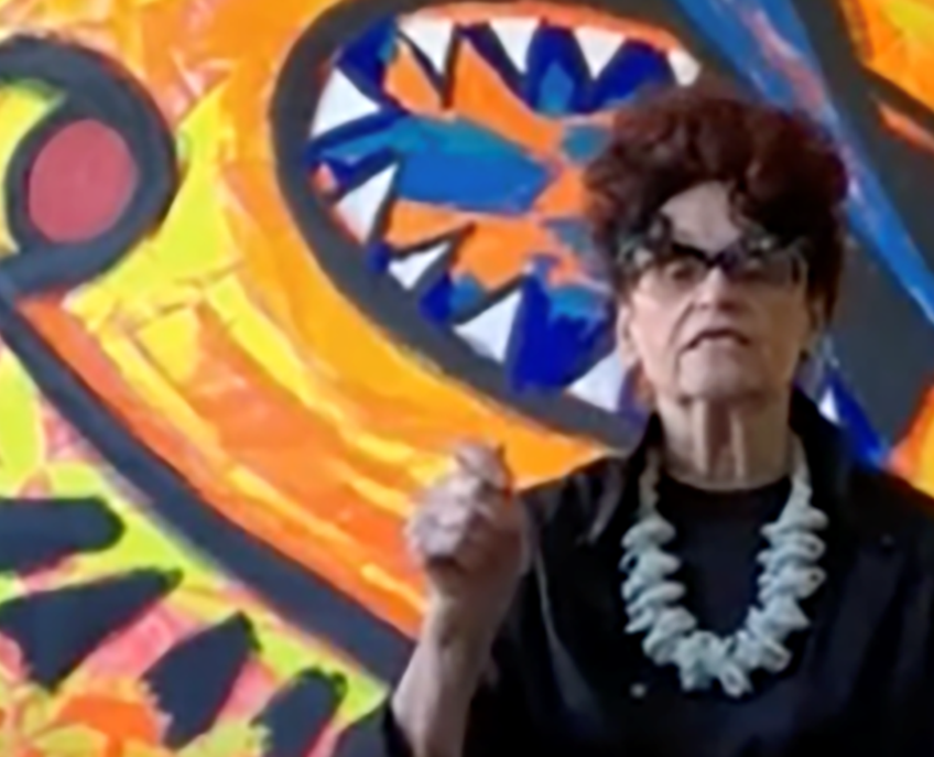 Detail of color screenshot of Judith Bernstein in front of one of her paintings wearing a black blouse and light-colored, chunky necklace holding her hand up in a fist as if making a point in conversation.