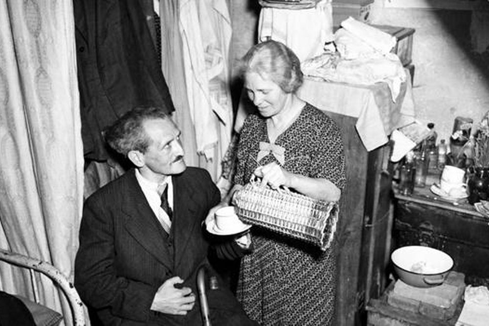 A white woman in a dress pours tea for a seated man in a suit, in a small home crowded with items (mobile)