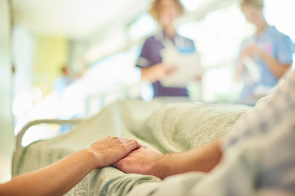 Close-up of person in hospital bed holding hands, with nurses blurred in the background