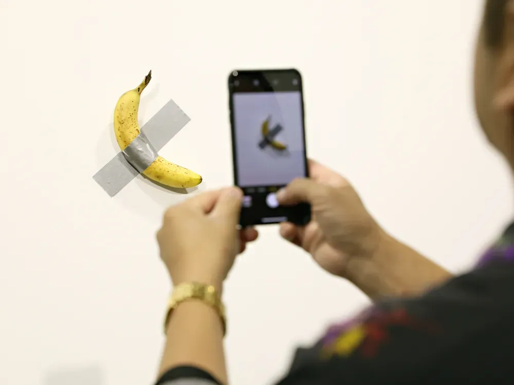 Person taking a photo of a banana taped to a white wall