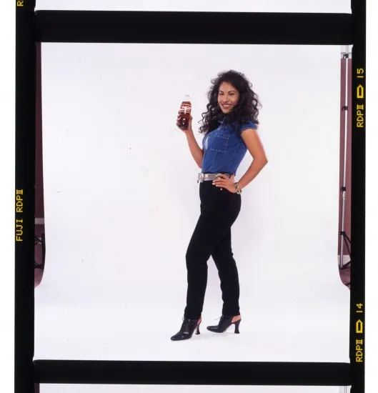 Selena in a blue vest and black pants holding a bottle of Coca-Cola