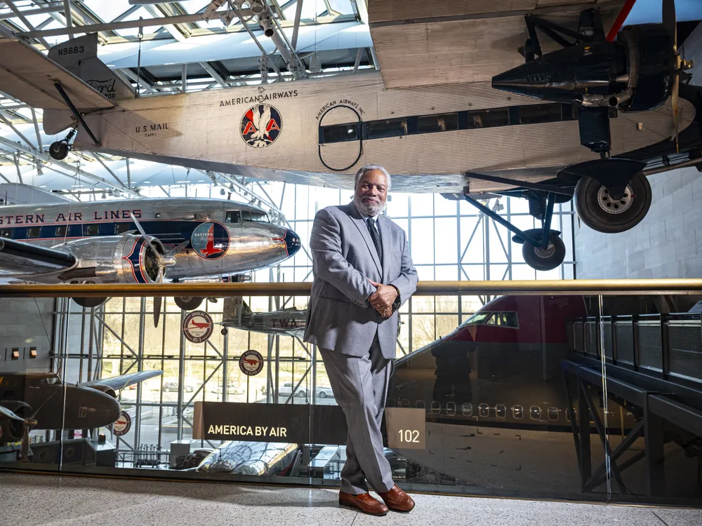 A Black man in his early 70s, wearing a blue suit and brown dress shoes, stands on a mezzanine overlooking a museum gallery displaying at least five airplanes.