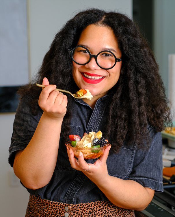 Image shows a smiling woman eating a bowl of fruit.