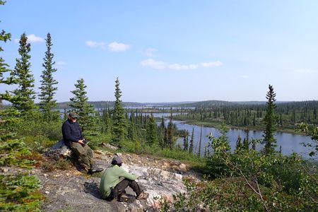 Two men sit on a rock bed on top of a forested hill over looking a remote lake.