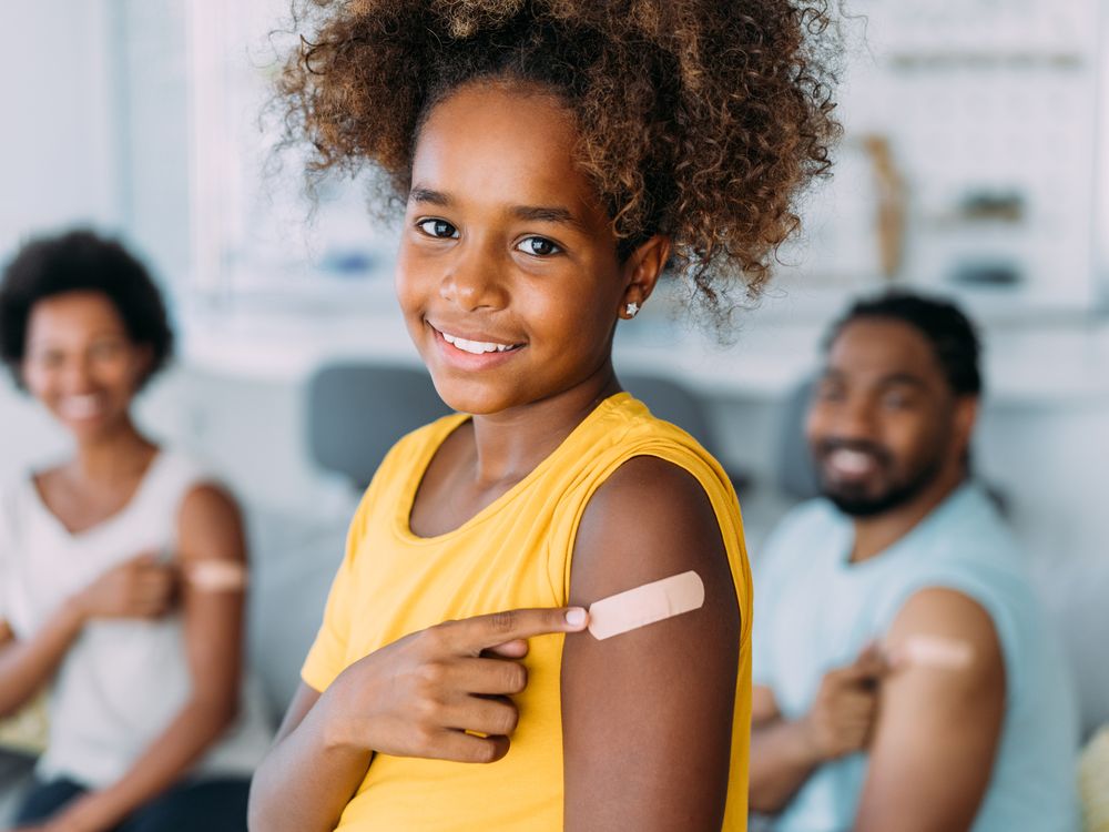 A young girl pointing to a bandaid on her left arm and smiling with parents in background