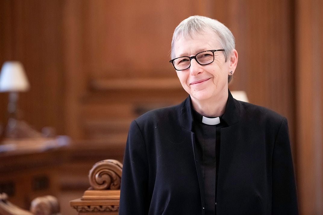 Woman in black jacket and white clerical collar, with black-rimmed glasses and short gray hair, smiles.